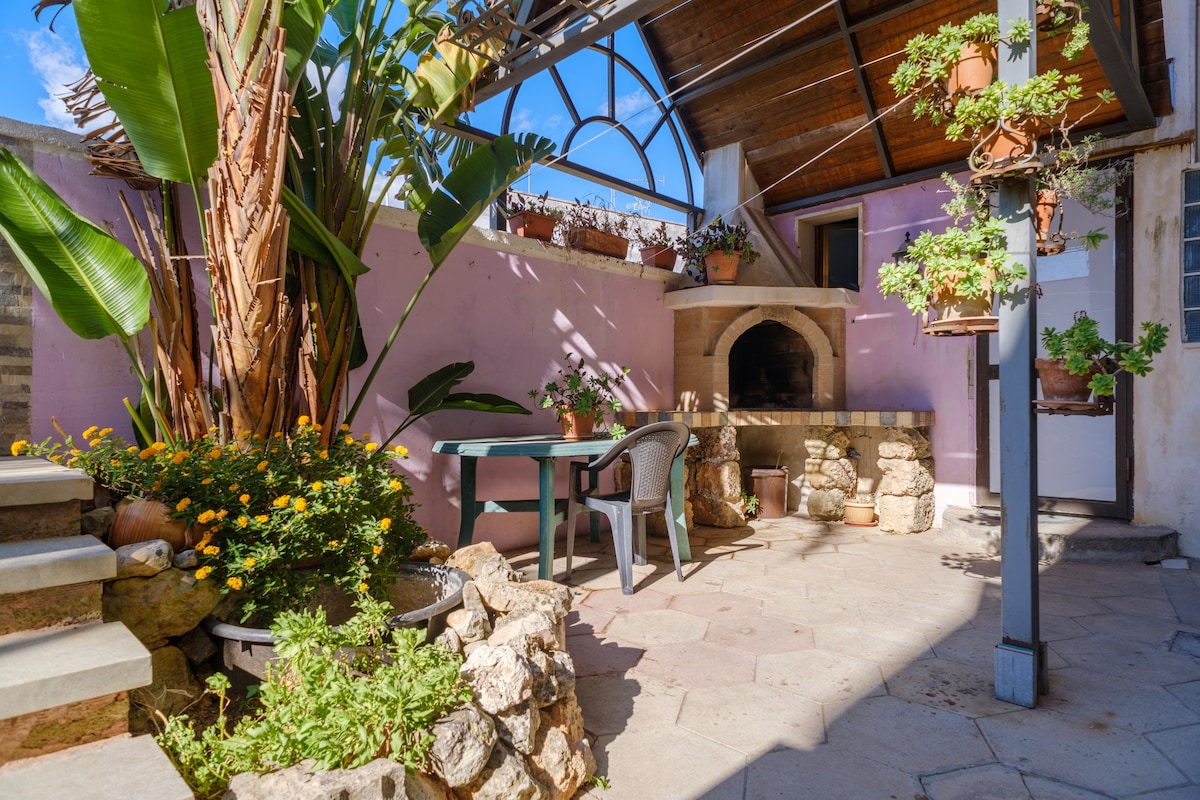 An outdoor area features a covered terrace with a barbecue and seating space. A stone wall is adorned with potted plants, alongside vibrant flowers and lush greenery. The solid surface is partially shaded, creating a relaxed environment for gatherings.