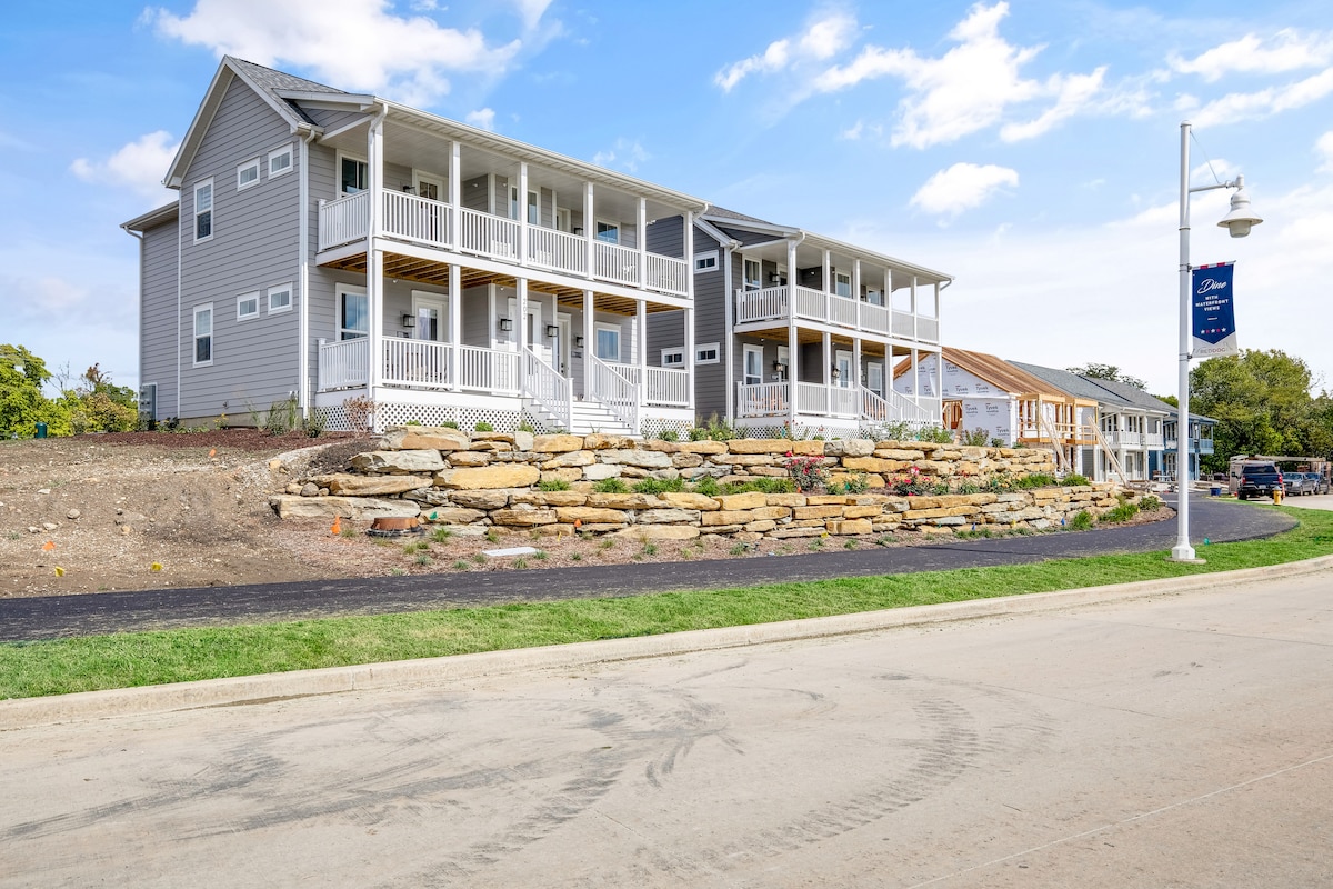 A charming multi-unit building features multiple balconies overlooking a landscaped area. Natural stone accents are visible in the front yard, while the structure showcases a combination of light grey siding and darker trim. A well-maintained path leads from the street to the entrance.