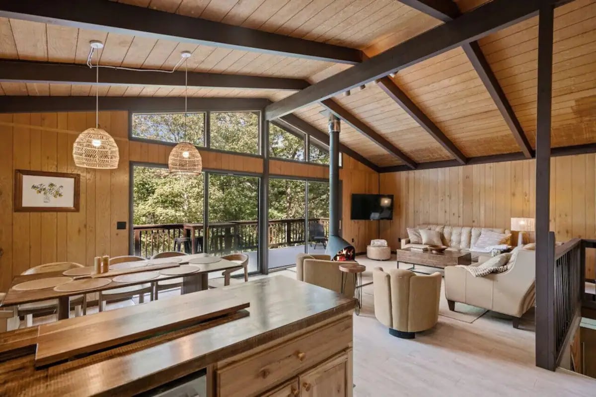 An open living space features a high, vaulted ceiling with wooden beams. Large windows provide ample natural light and views of nature. A cozy seating area includes a sectional sofa and armchairs, while a dining table is located nearby. A wood-burning stove is positioned against a wooden wall.