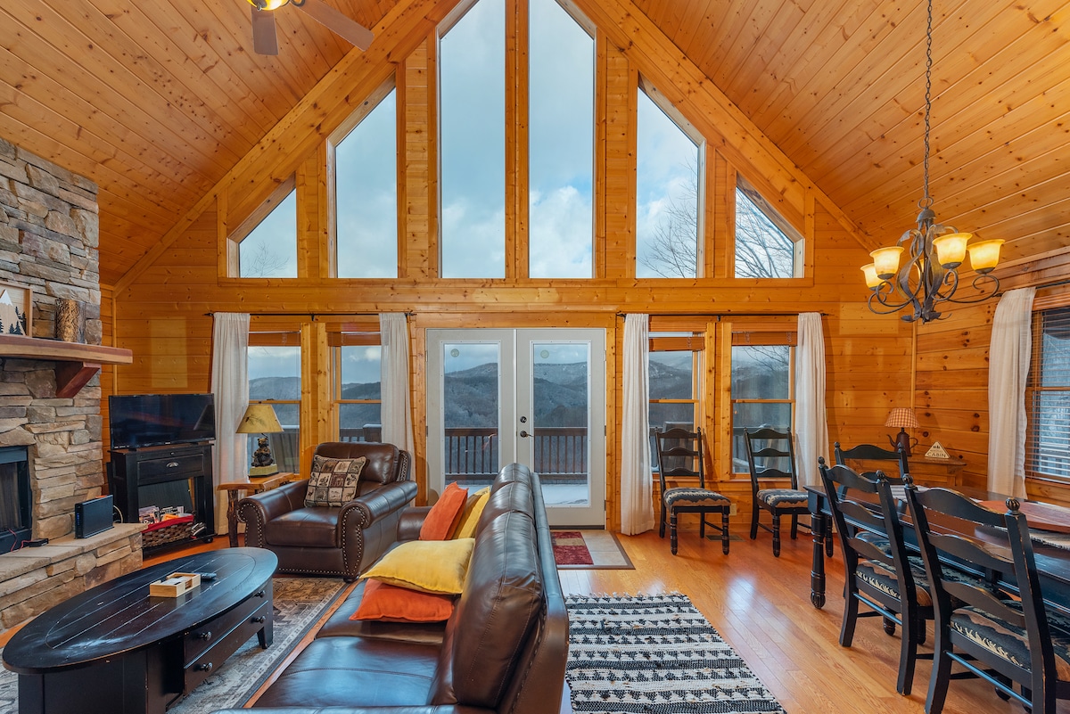 A spacious living area features high ceilings with large windows, allowing natural light to illuminate the wooden interior. Comfortable seating arrangements are visible, along with a dining area equipped with a wooden table and chairs. A stone fireplace adds a rustic touch.