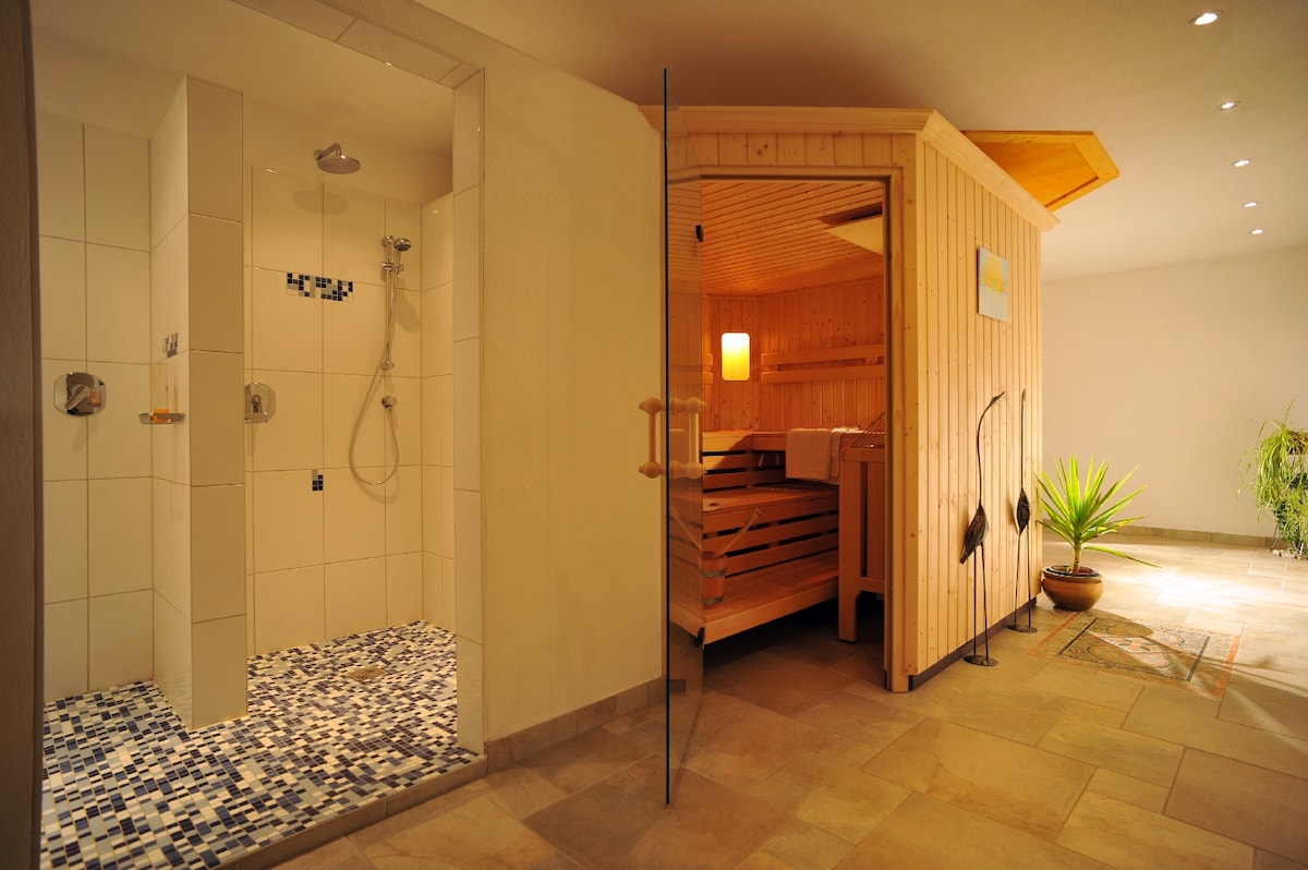 A serene wellness area features a wooden sauna room adjacent to a shower space. The sauna showcases warm wooden tones, while the shower area is defined by clean, tiled walls and a mosaic floor. A potted plant adds a touch of nature to the inviting environment.