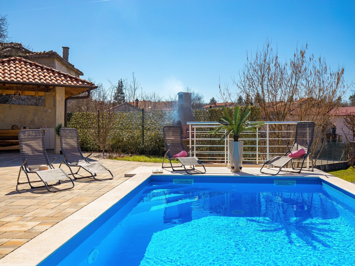 A heated outdoor swimming pool is surrounded by a paved deck, featuring four reclining lounge chairs positioned for relaxation. Lush greenery and a garden area are visible in the background, along with a covered outdoor seating area, creating an inviting space for leisure.