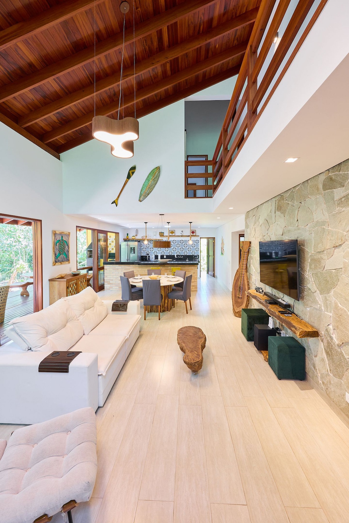 A spacious living area features a white sofa and a natural wood coffee table, complemented by stone accents on the wall. High ceilings with wooden beams create an open feel. The dining area, visible in the background, includes a table set for six, with views of the outdoors.