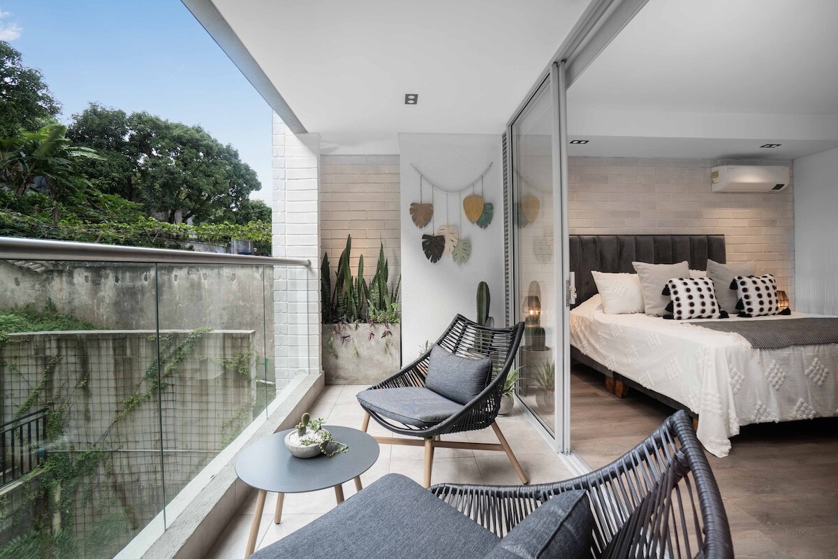 A cozy balcony area is showcased with two wicker chairs and a small table, providing a serene space to relax. The glass doors open to a well-lit bedroom in the background, decorated with neutral tones and an inviting bed framed by green plants. Views of the surrounding garden are visible.