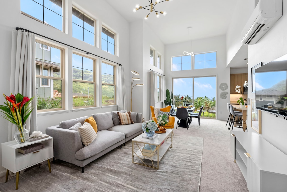 A bright living room showcases large windows allowing natural light and views of the landscape. A comfortable gray sofa is adorned with decorative pillows, while a coffee table holds a vase of flowers. The room flows into a dining area with a view, creating an inviting space.