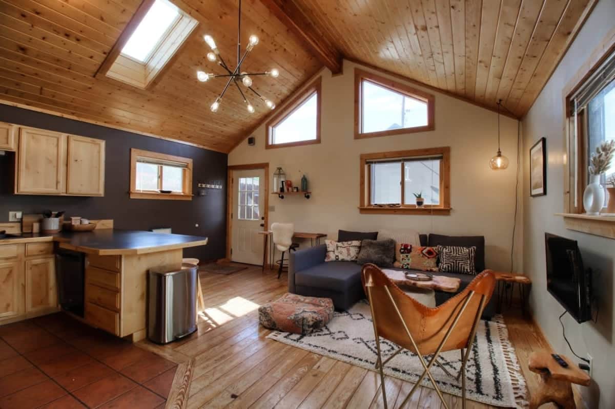 A bright living area features high wooden ceilings and a cozy seating arrangement. Large windows allow natural light, while a unique light fixture adds character. The space includes a kitchenette with modern appliances, and a small table, creating an inviting space for relaxation.