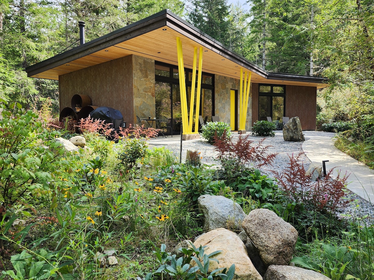The Shed is surrounded by lush greenery and colorful flowers, with a modern exterior that features a mix of stone and rust-colored siding. Playful yellow supports contrast with the natural setting, while large windows provide views of the serene forest landscape.