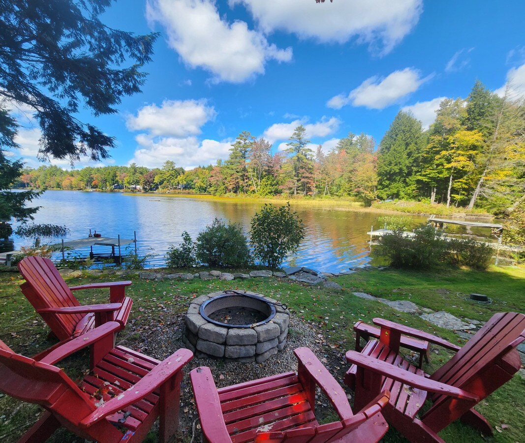 A serene outdoor space features four red Adirondack chairs arranged around a stone fire pit, overlooking a tranquil lake. Lush greenery and colorful trees create a natural backdrop, while a boat dock is visible along the water's edge under a clear blue sky.