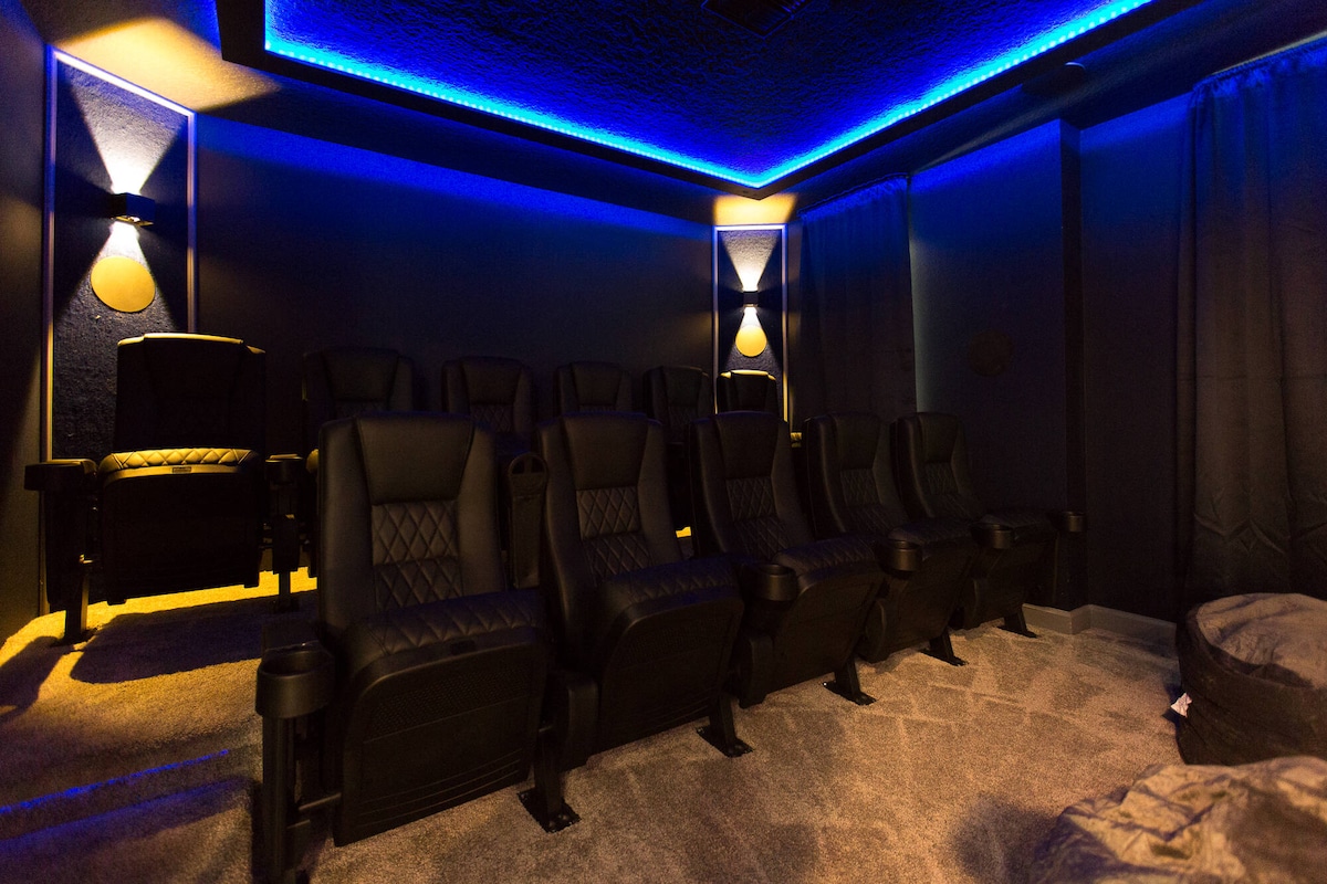 A cinema room is featured with plush seating arranged for 11, illuminated by soft blue LED lights along the ceiling. Dark curtains frame the windows, and carpeted flooring contributes to a cozy ambiance suitable for movie nights.