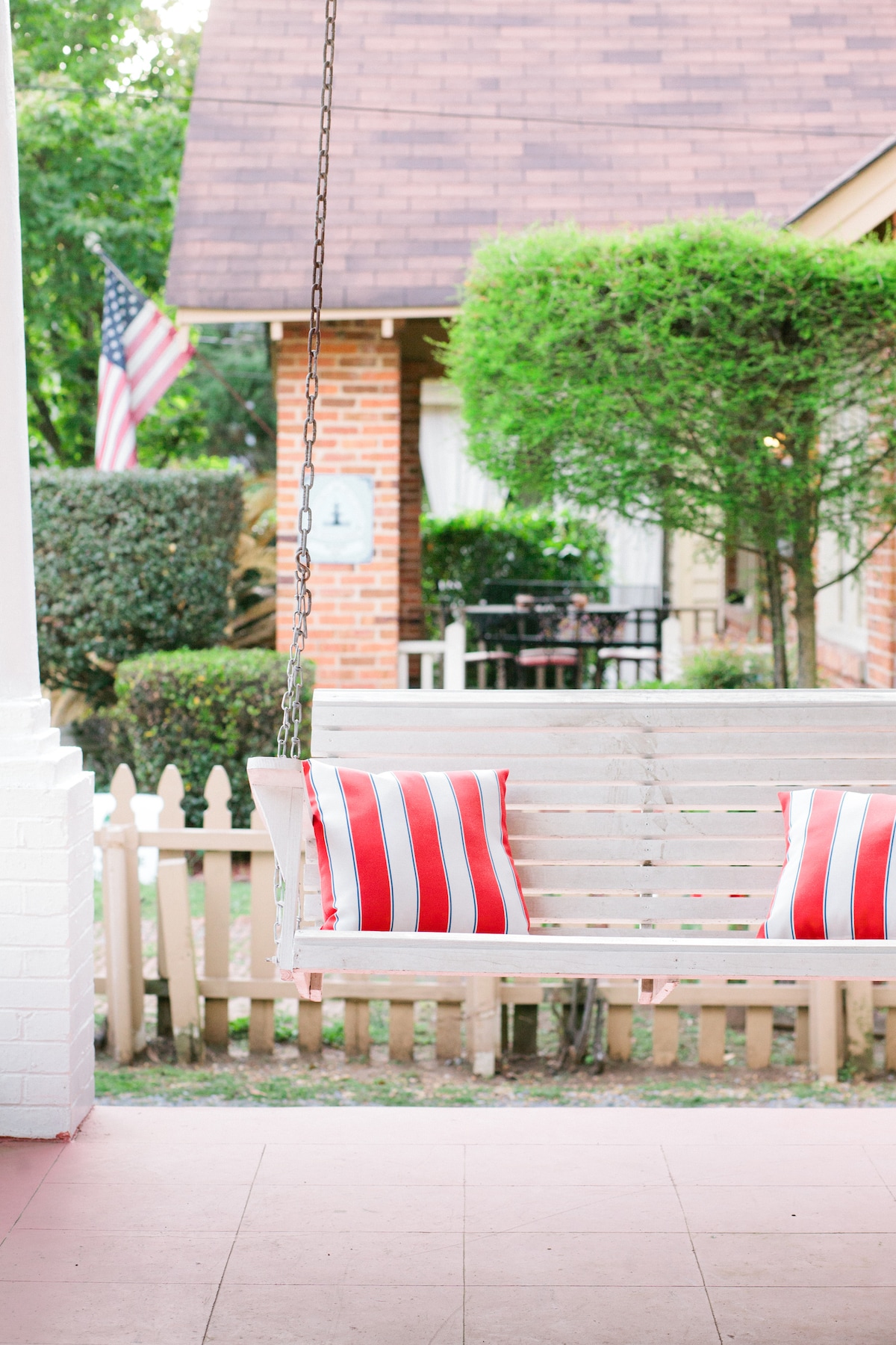 A wooden swing adorned with red and white striped pillows is suspended from above, offering a spot for relaxation. In the background, a brick home is visible, along with a neatly maintained garden, white picket fence, and an American flag gently waving.