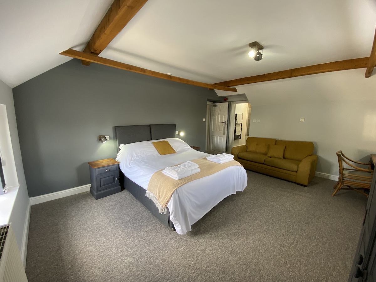 A spacious family room features a large bed covered with white linens and a tan throw blanket. A cozy sofa is positioned against one wall, and natural light fills the room through a window. Exposed wooden beams add a touch of character.