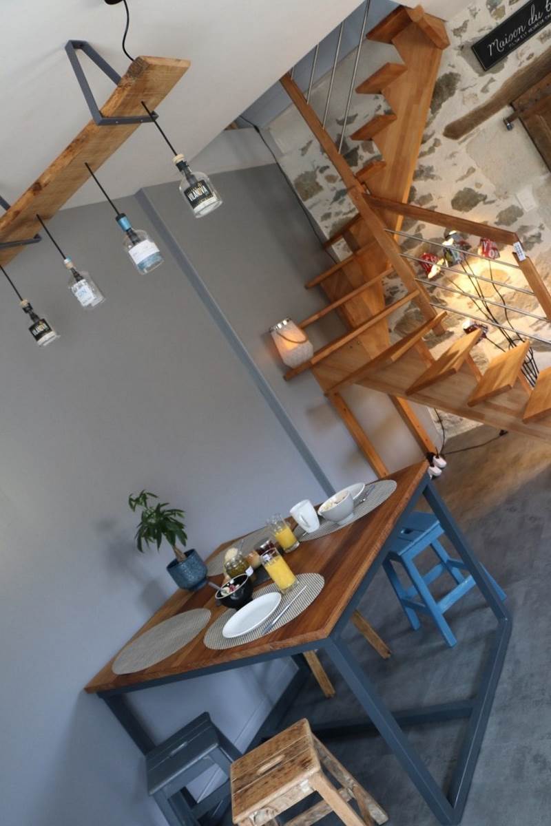 A wooden dining table is set for breakfast with plates, cutlery, and beverages, positioned under modern pendant lights. A small plant adds a touch of greenery. In the background, a wooden staircase with a simple design leads to the upper level, enhancing the cozy ambiance.