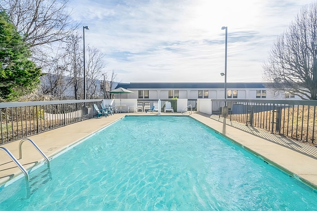 An outdoor swimming pool is surrounded by a concrete patio. Loungers and umbrellas are positioned along the pool's edge. The water appears clear and inviting, with a low fence providing safety. A building is visible in the background, featuring large windows.
