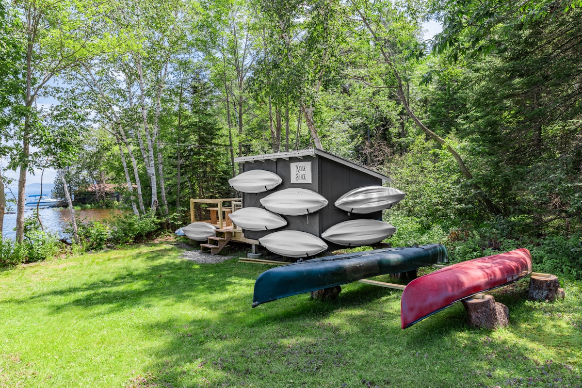 A grassy area beside a forested background features a storage shed displaying multiple white canoes on its side. Two larger canoes sit on the ground; one is red and the other is dark blue. A wooden deck can be seen adjacent to the shed, providing a space for relaxation.
