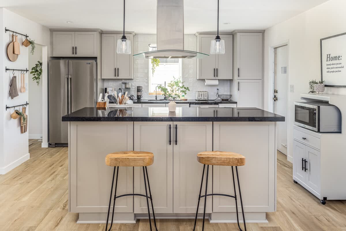 A modern kitchen features a large island with two wooden stools. Sleek cabinetry and stainless steel appliances complement the dark countertops. Soft lighting from pendant fixtures creates a cozy ambiance, while ample natural light streams through the window, enhancing the inviting workspace.
