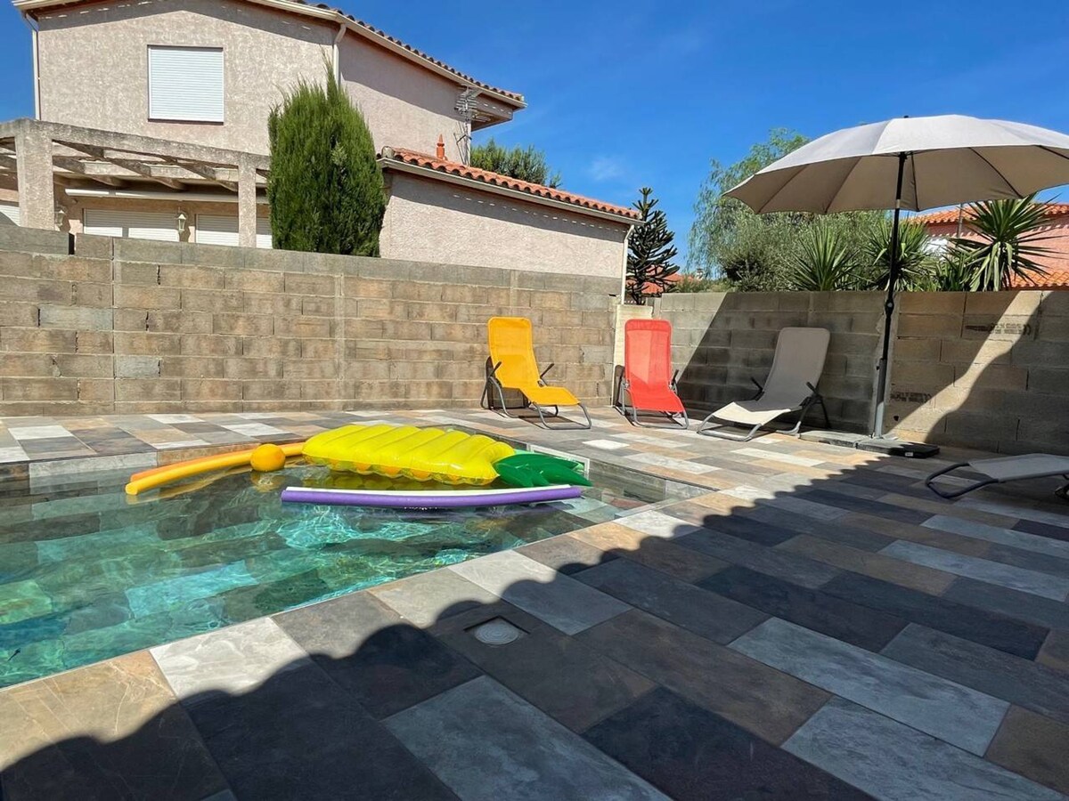 A private swimming pool features a colorful inflatable float and is surrounded by tiled decking. Two yellow deck chairs and two lounge chairs provide seating under a large umbrella, with greenery visible in the background. The area is sheltered, offering a calm atmosphere.