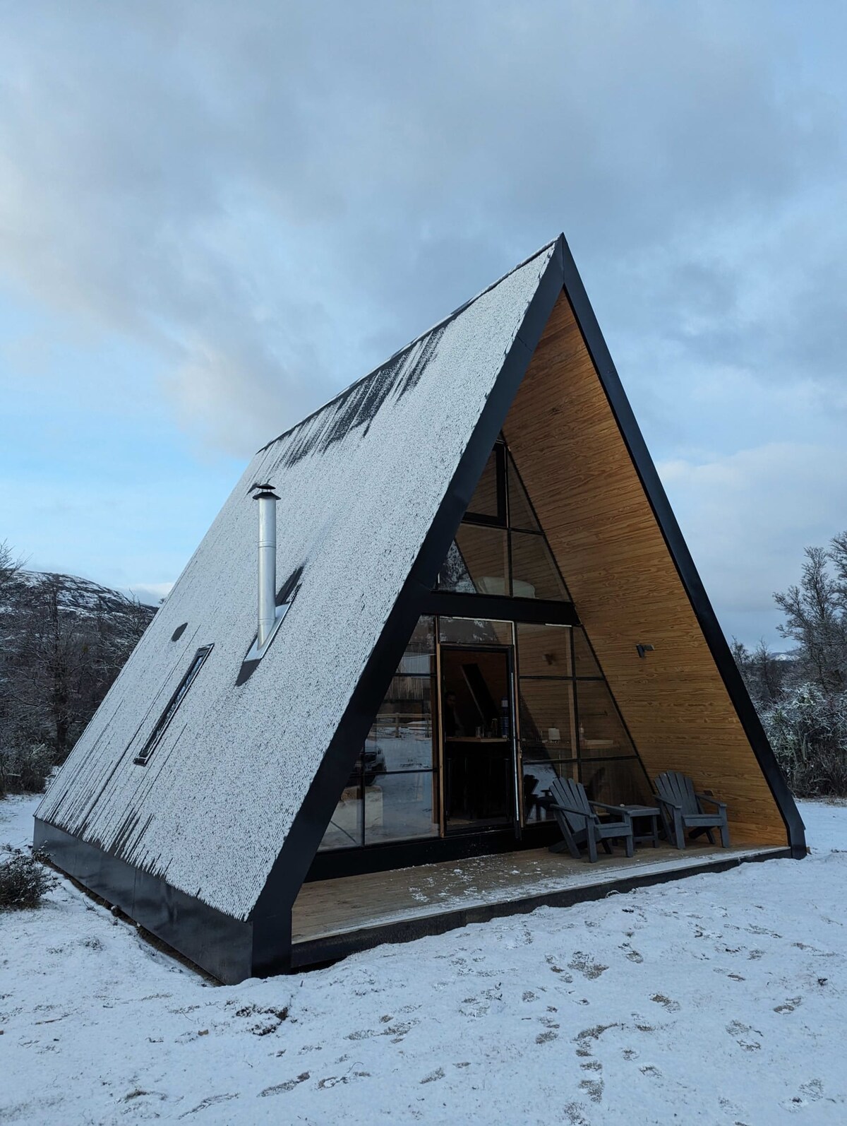 An A-frame cabin is set in a snowy landscape, with a wooden deck featuring two adirondack chairs. The structure is accented by large glass windows and a chimney, blending modern design with a natural setting.