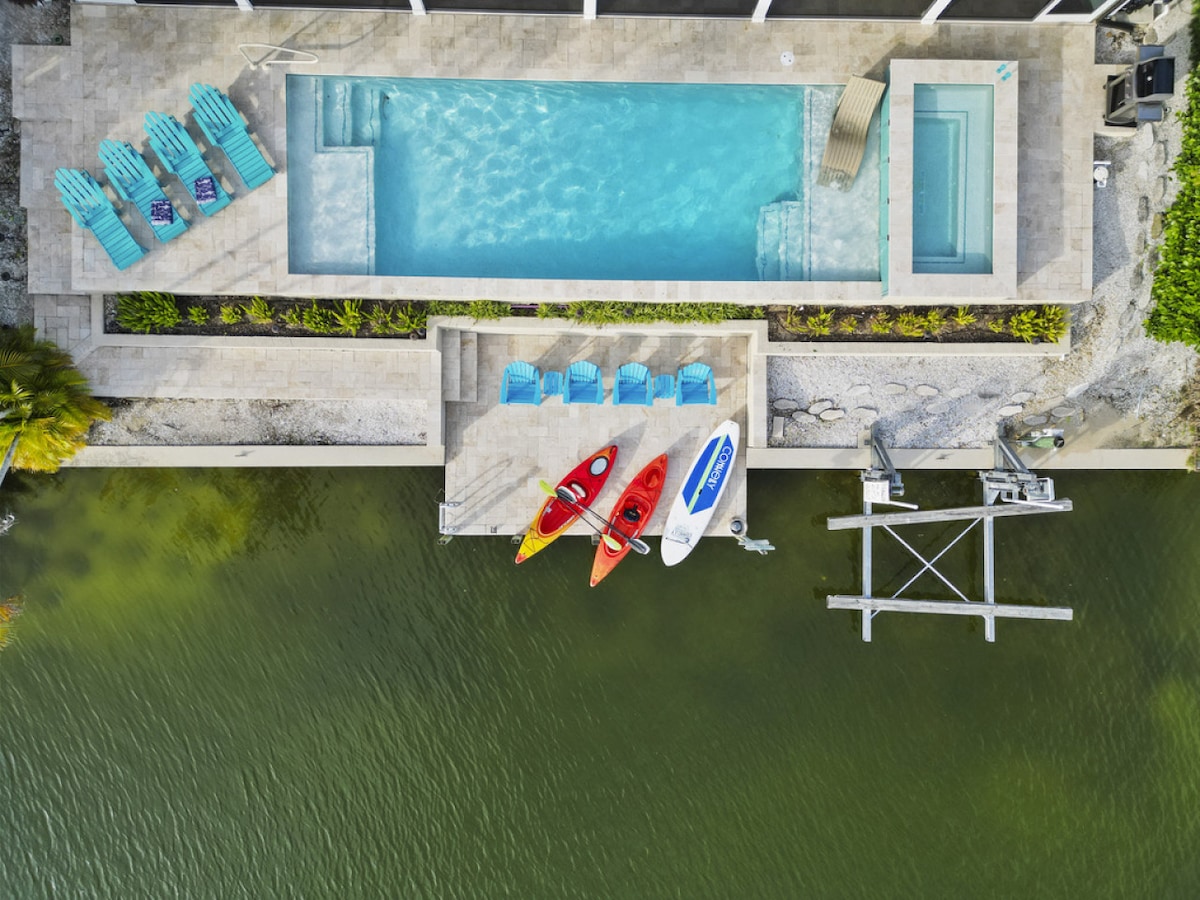 An aerial view captures the heated saltwater pool alongside a spacious deck. Colorful kayaks and paddleboards are arranged on the dock, which offers direct access to the serene lake. Lounge chairs provide comfortable seating in the sun, surrounded by well-maintained landscaping.
