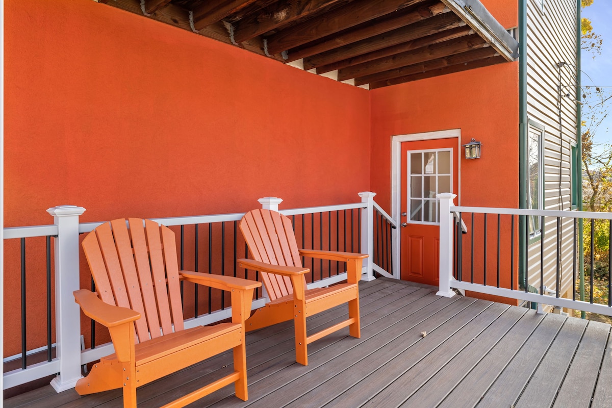 A bright orange wall complements the wooden deck, where two orange chairs are positioned for outdoor seating. The entrance door features a large glass panel, flanked by a light fixture, enhancing the welcoming ambiance.