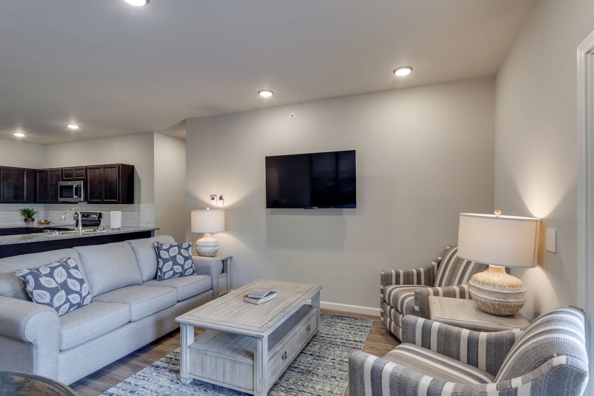 A modern living area features a light gray sofa with decorative pillows and two striped armchairs. A wooden coffee table sits in the center, complemented by a patterned area rug. A flat-screen TV is mounted on the wall, with soft lighting illuminating the space.