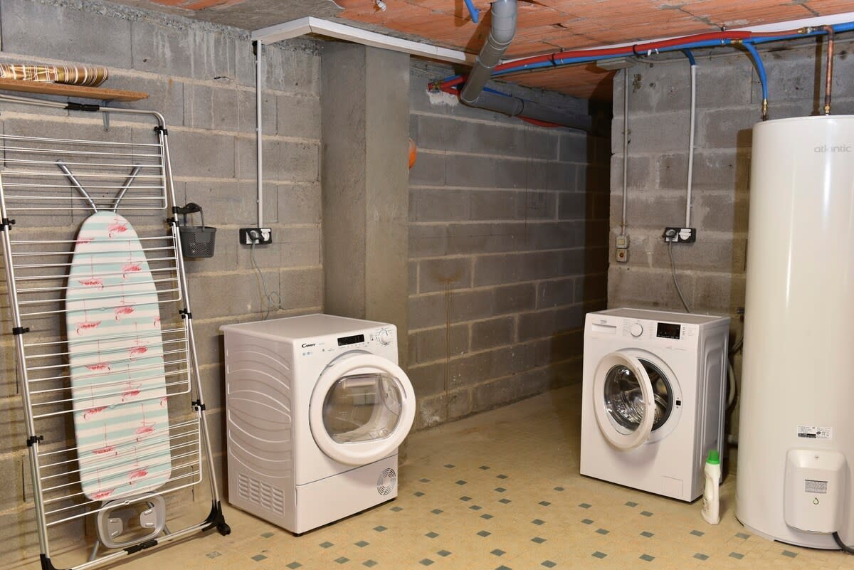 A functional laundry area features a drying rack, a washing machine, and a dryer. The appliances are positioned against a concrete wall, with utility connections visible. The floor is covered in a simple tile pattern.