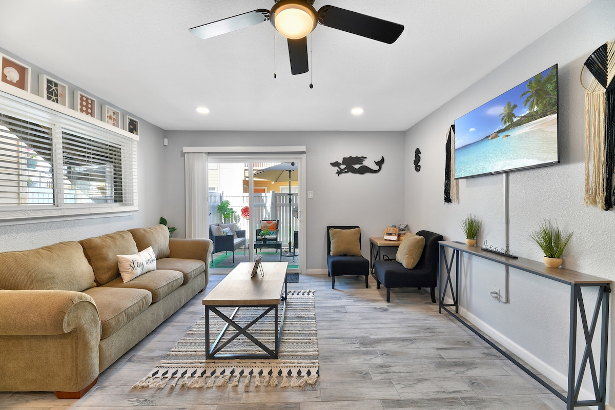 A spacious living room is furnished with a comfortable sofa and armchair, complemented by a coffee table and a flat-screen TV. Natural light enters through large windows, with sliding doors leading to a private patio. Decorative accents include wall art and a patterned area rug.