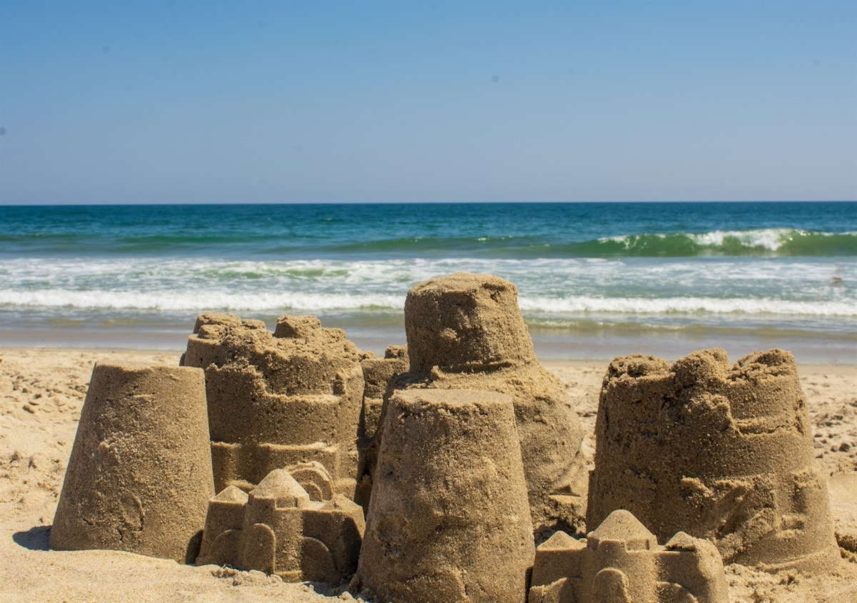 A collection of sandcastles sits along the shoreline, varying in height and design. Waves gently lap at the sandy beach, while a clear blue sky stretches overhead, enhancing the coastal ambiance.