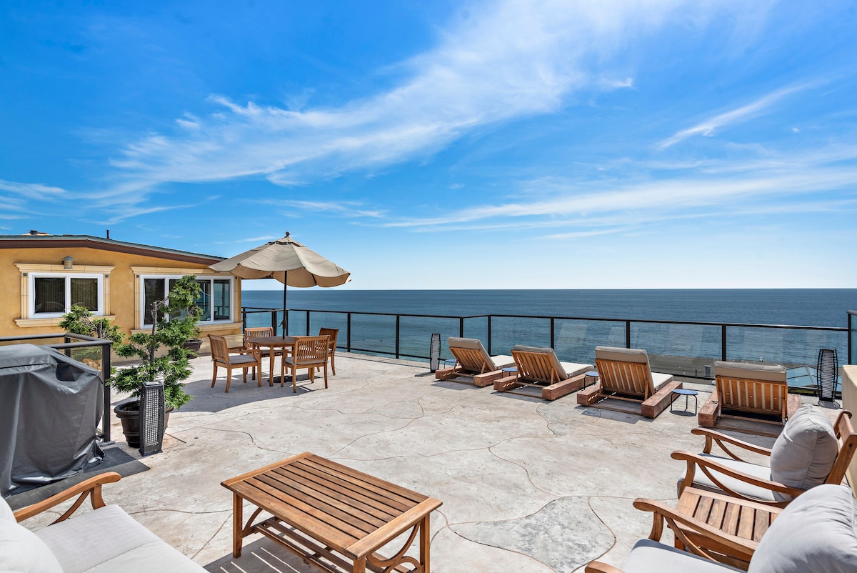 A spacious rooftop patio is featured, lined with wooden lounge chairs and a dining table. An umbrella provides shade, while a barbecue grill stands nearby. The expansive ocean views are framed by a clear blue sky, creating an inviting outdoor space for relaxation.