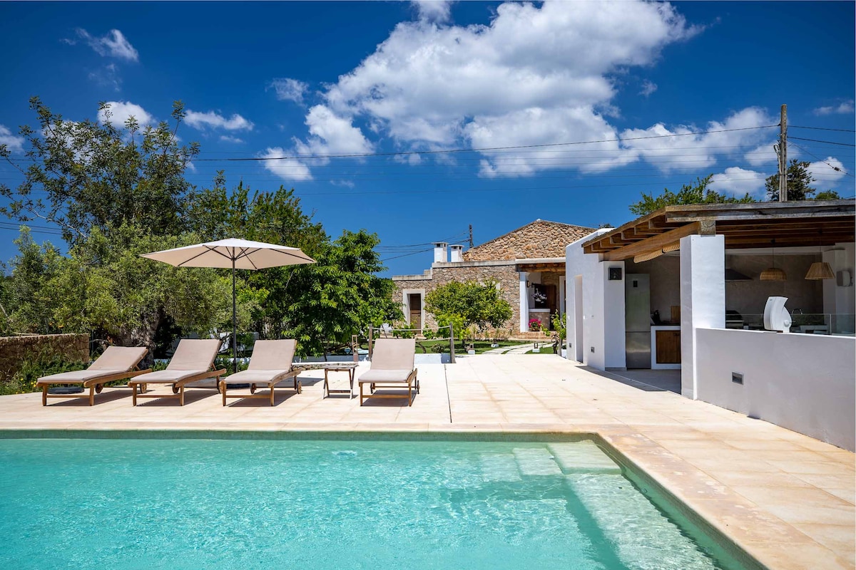 A serene outdoor area is showcased, featuring a clear swimming pool bordered by sun loungers. An umbrella provides shade, while an outdoor kitchen and dining space are visible in the background. Lush greenery complements the tranquil atmosphere under a bright blue sky.