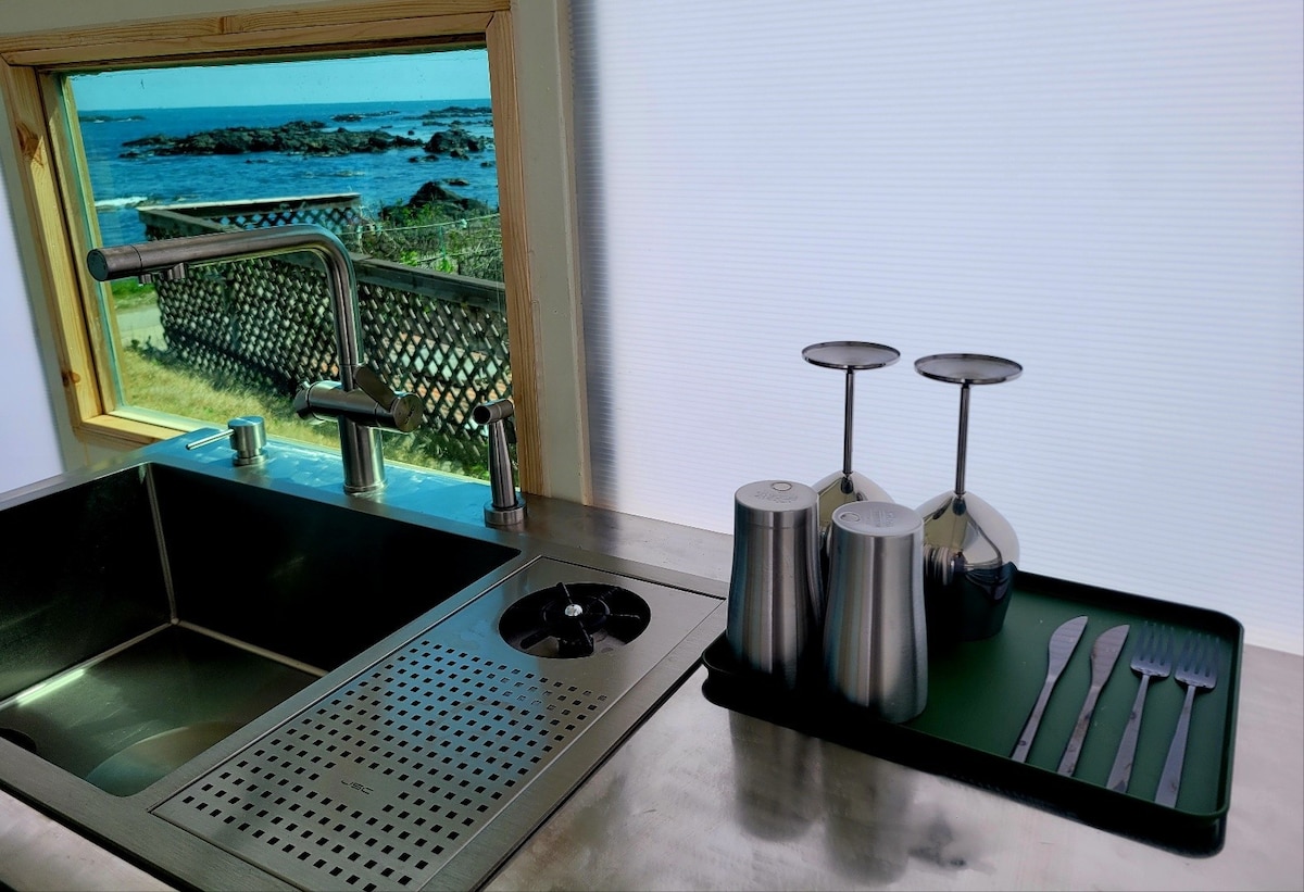 A well-equipped kitchen area is shown, featuring a stainless steel sink and dish rack with glasses. A window reveals a scenic view of the ocean and surrounding greenery, allowing natural light to enter the space.