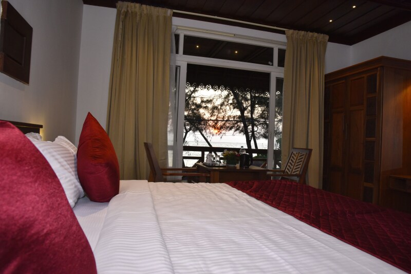 A spacious bedroom features a large bed with crisp white linens and red accent pillows. Natural light fills the room from a large window, showcasing a sunset over the water. Wooden furniture, including a table and chairs, is positioned near the window.