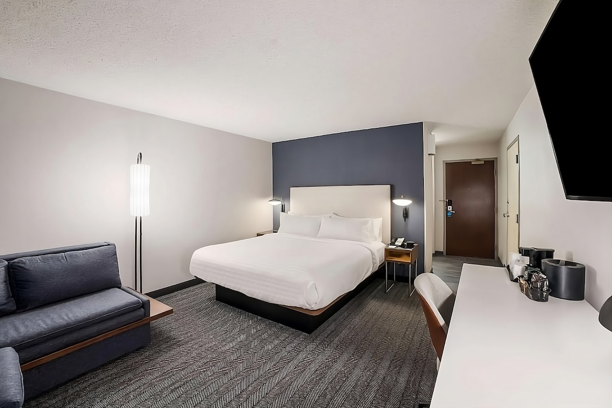 A spacious hotel room features a king bed with white linens, complemented by a dark accent wall. A comfortable sofa and a sleek work desk with an ergonomic chair are included. A flat-screen TV and a doorway lead to the exterior, enhancing accessibility.