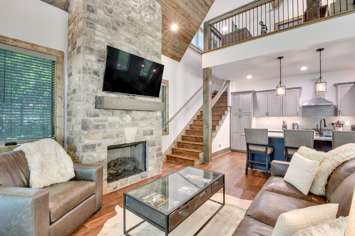 A spacious living area features a stone fireplace and large windows allowing natural light. Comfortable leather seating is arranged around a glass-top coffee table, with a view of the open kitchen and dining area in the background, showcasing modern appliances and bar seating.