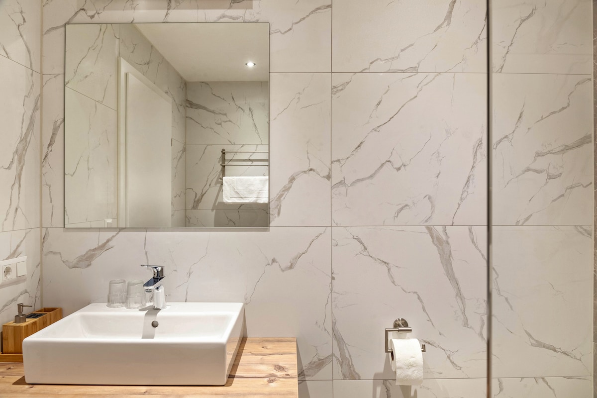 A modern bathroom is showcased with marble-inspired wall tiles. A large mirror is mounted above a rectangular sink, positioned on a wooden countertop. A towel rack is visible in the background, and essential toiletries are neatly arranged nearby.