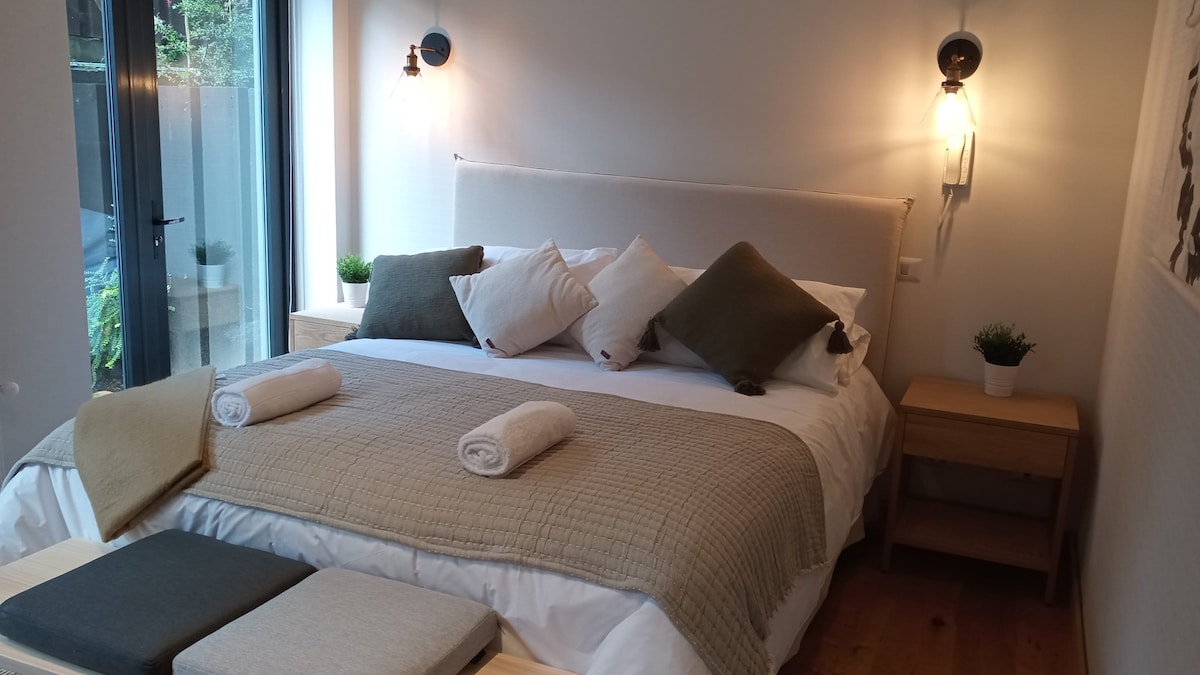 A spacious bedroom is presented, featuring a large bed with an array of pillows. Soft, neutral-toned bedding covers the bed, complemented by rolled towels on the edge. Natural light enters through floor-to-ceiling windows, accented by two stylish wall sconces.