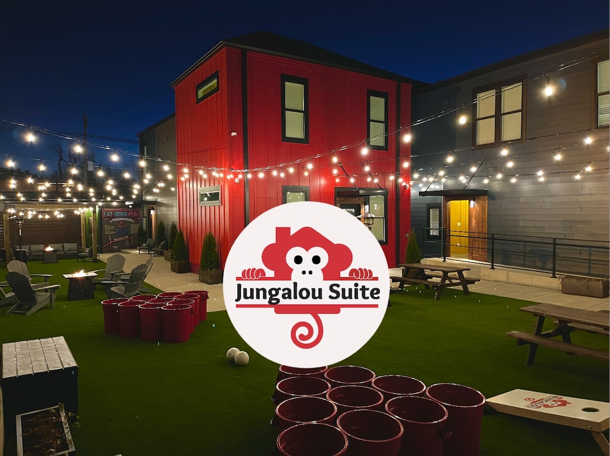 The vibrant exterior of the Jungalou Suite is displayed at night, highlighted by warm string lights overhead. A grassy game area features large red cups and bean bag toss games, while the surrounding buildings offer an inviting urban atmosphere.
