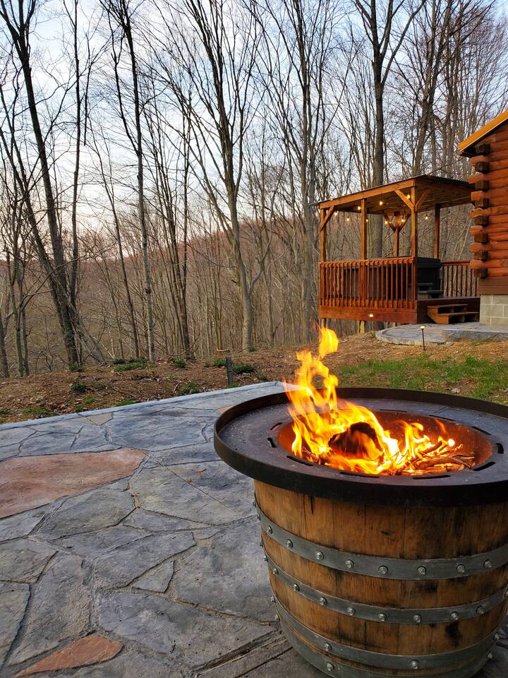 A warm fire burns in a rustic barrel-shaped fire pit on a stone patio, surrounded by natural scenery. In the background, a wooden deck is visible, offering a serene view of the wooded landscape as the sun sets behind the trees.