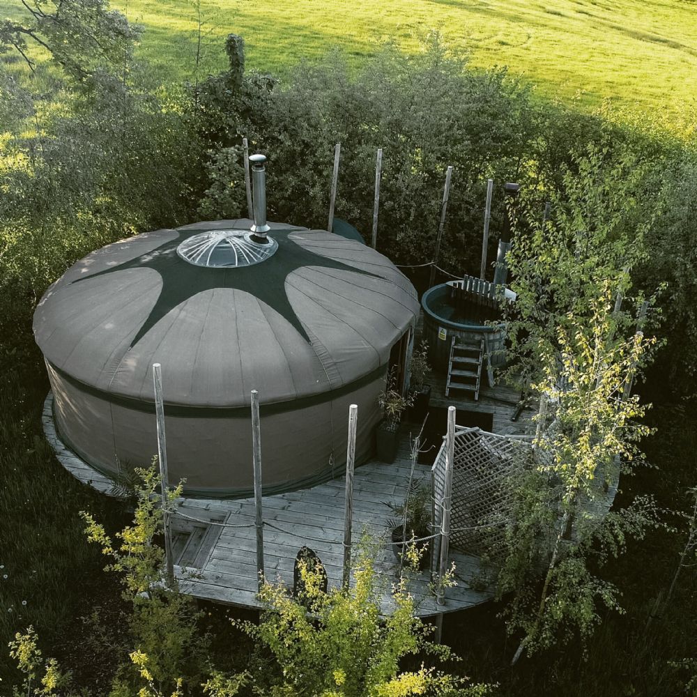 A circular yurt is positioned on a wooden deck surrounded by lush greenery. A hot tub is located nearby, with a wooden ladder leading up to it. The yurt features a central skylight that allows natural light to illuminate the interior.