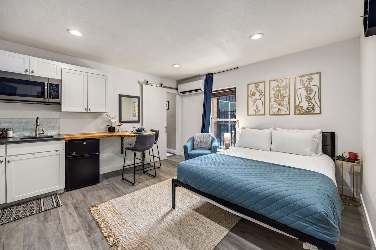 The studio apartment showcases a cozy sleeping area with a spacious bed covered in a blue quilt. A kitchenette with sink and refrigerator is located to the left, alongside a bar table with seating. Natural light enters through the window, illuminating the neutral decor.