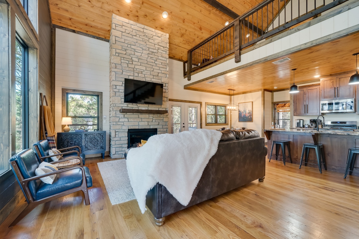 A spacious living area features a stone fireplace and a large flat-screen TV mounted above. A plush dark sofa, layered with a textured throw, faces the fireplace, while a light area rug anchors the seating. Natural light fills the space through large windows, enhancing the wooden ceiling.