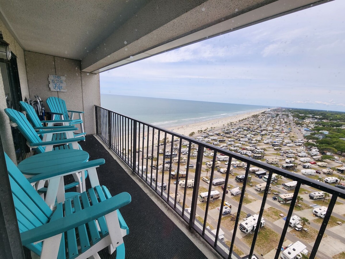 A spacious balcony offers a view of the Atlantic Ocean and the coastline stretching into the distance. Four bright blue adirondack chairs are arranged along the railing, inviting relaxation while enjoying the scenic beach landscape below.