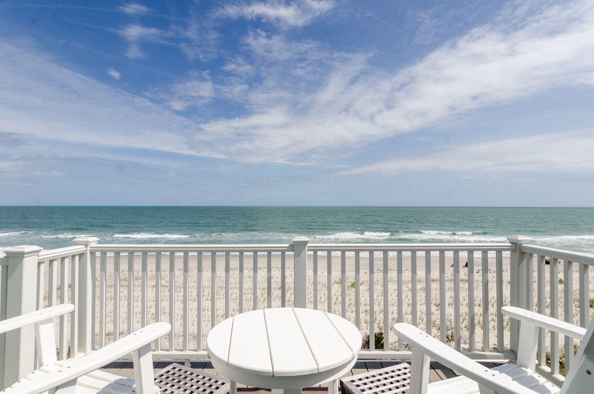 A spacious deck overlooks the ocean, featuring white wooden railings and a circular table surrounded by two chairs. The picturesque view encompasses sandy beach and gentle waves beneath a clear blue sky, inviting relaxation and enjoyment of the coastal scenery.