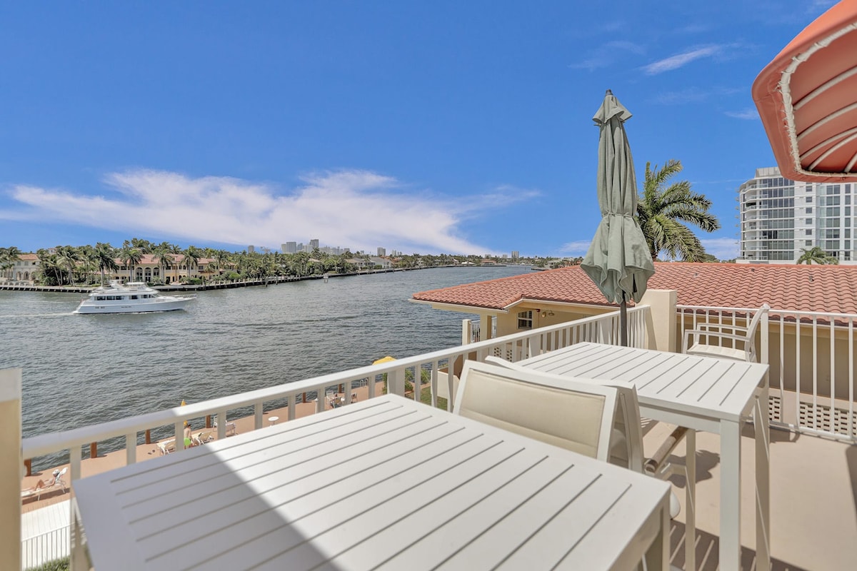 An outdoor patio features white dining tables with umbrellas, overlooking the Intracoastal waterway. The calm water reflects the clear blue sky, while palm trees provide a serene backdrop. Nearby, a boat glides along the water, enhancing the tranquil ambiance of the setting.