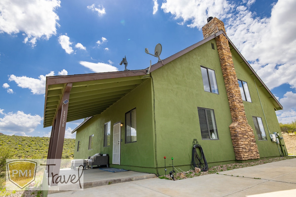 The exterior of the home features a two-story structure with green walls and a stone chimney. A covered patio is visible, along with large windows that allow natural light inside. The driveway leads to the entrance, surrounded by scenic hills.