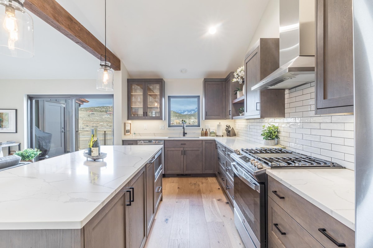 A modern kitchen features dark cabinetry and white countertops, enhanced by natural light from large windows. High-end appliances, including a gas range, are visible. Decorative elements, such as a plant and kitchenware, add a welcoming touch to the space connected to a dining area.