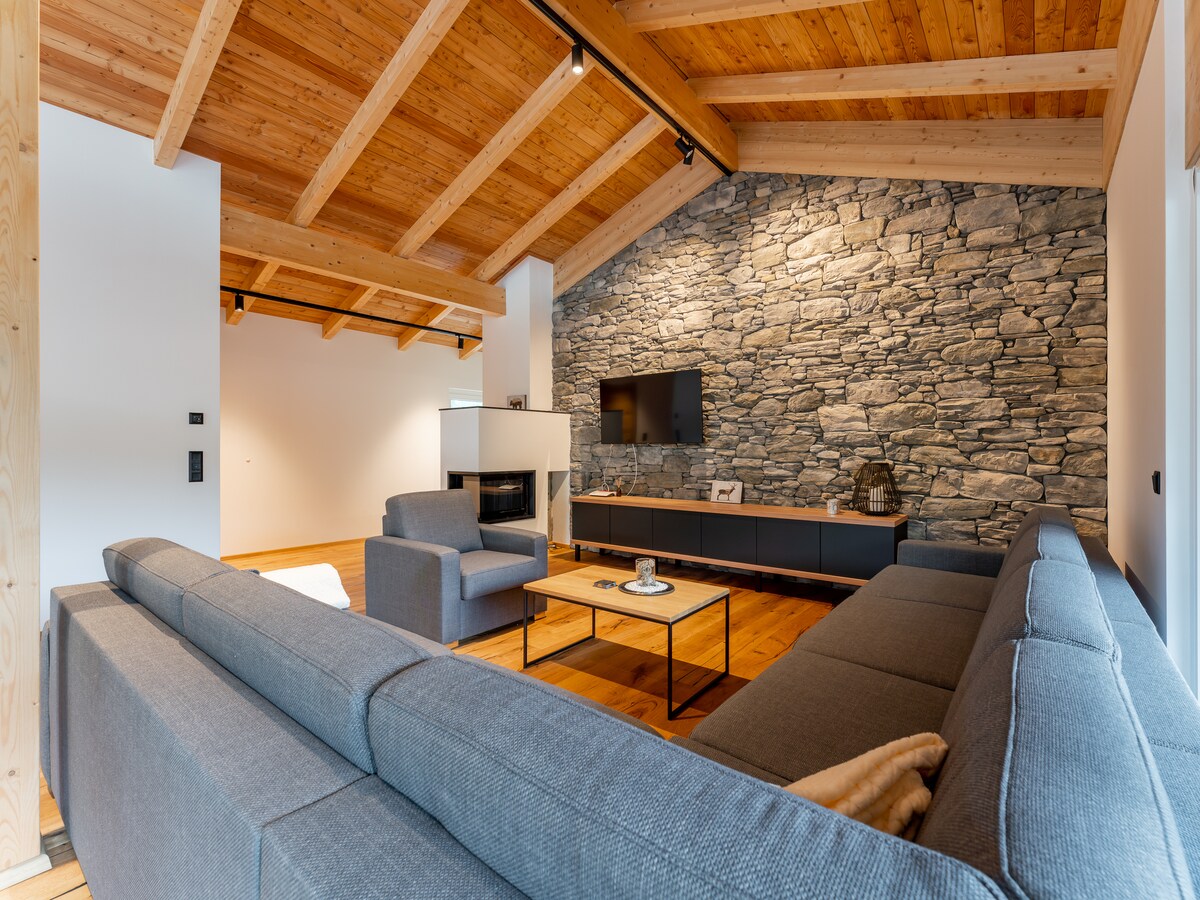 A spacious living area features a high wooden ceiling with exposed beams, enhancing the sense of openness. A stone accent wall serves as a backdrop for a wall-mounted TV, while a gray sectional sofa surrounds a central coffee table, creating an inviting space for relaxation.