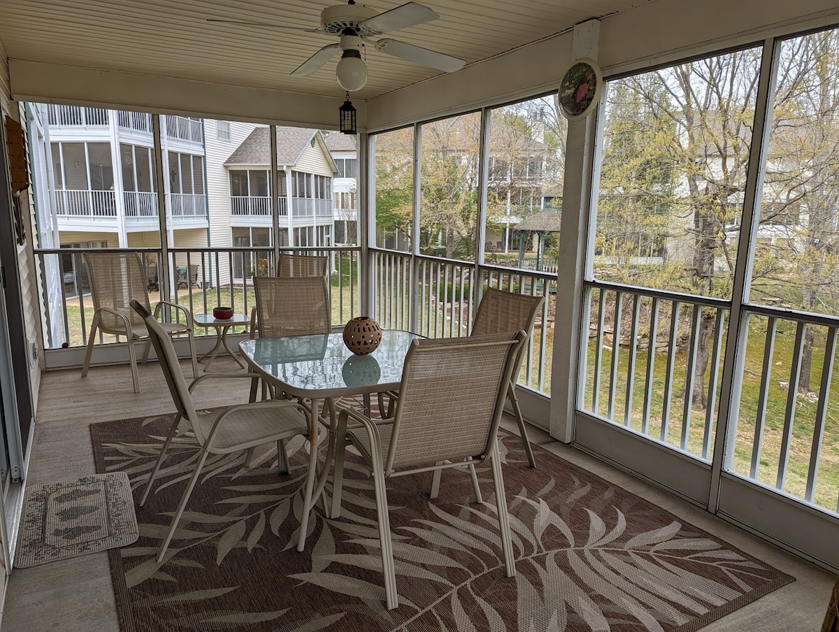 A screened-in porch features a glass dining table surrounded by four beige chairs, with a decorative bowl at the center. A patterned area rug adds texture to the floor. Surrounding green grass and trees can be seen through the mesh screens.