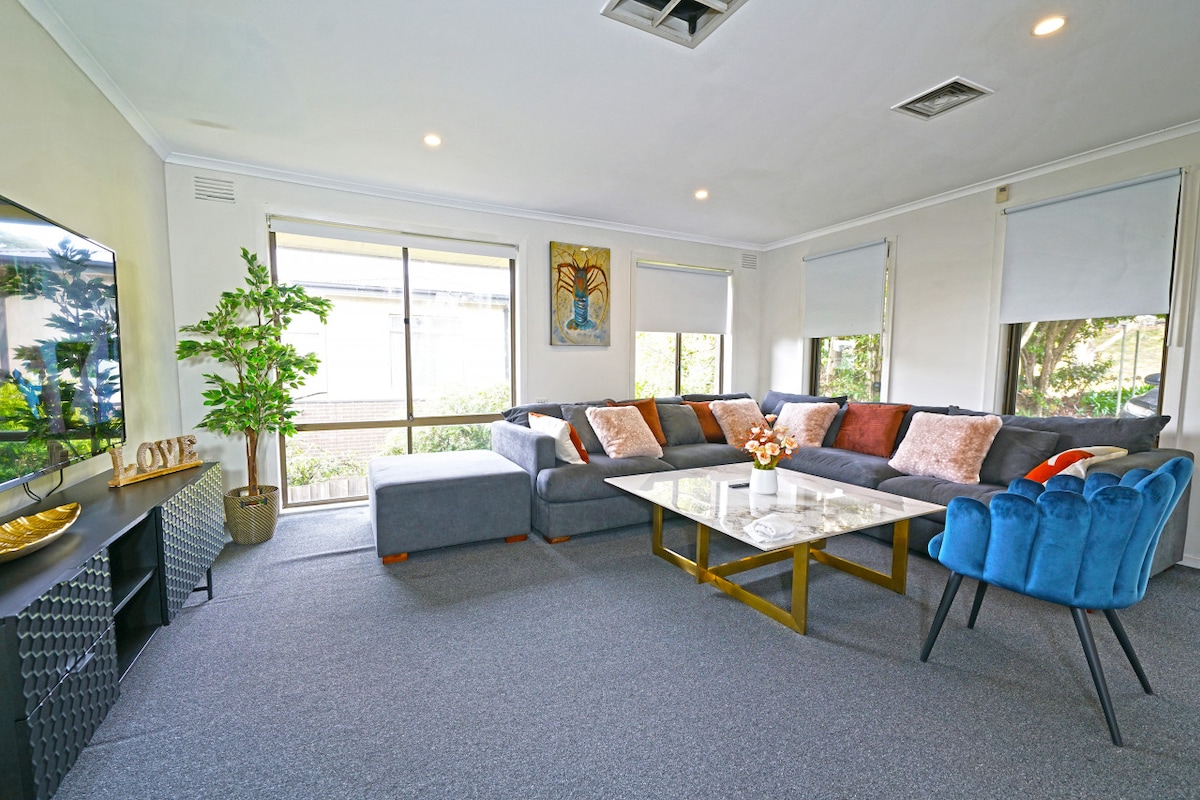A spacious living area features a large sectional sofa adorned with pillows, complemented by a blue accent chair. A glass coffee table sits in the center, and natural light filters through large windows, providing a view of the outside greenery.