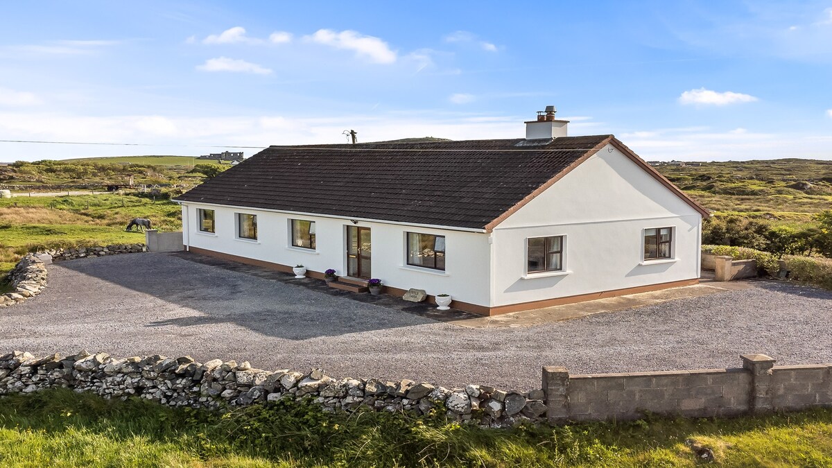 The exterior of a spacious bungalow is shown, featuring a white facade with a dark roof. A gravel driveway leads up to the front entrance. Surrounding the property, a stone wall and greenery create a natural setting, with several windows providing views of the landscape.
