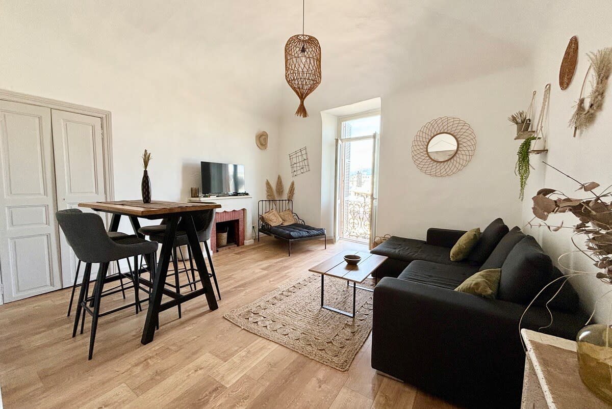 The bright living room features a large window offering views of the surroundings, with a comfortable sofa and a dining table with high stools. A TV is mounted on the wall, and decorative items are placed throughout, adding character and interest to the space.